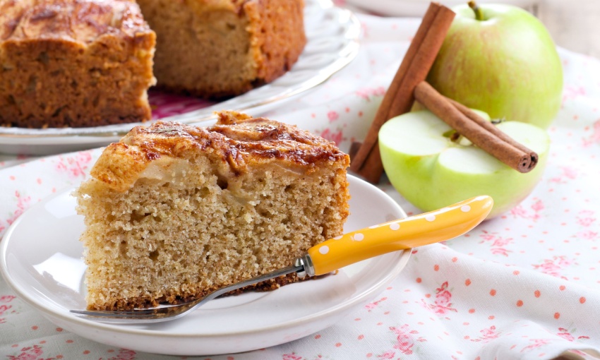 Fetta di torta di mele su un piatto, con accanto una posata. Sulla sfondo, delle mele verdi